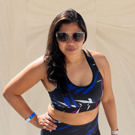 Woman wearing a colorful sports bra and leggings with sunglasses against a beige wall.